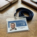A clear plastic ID badge holder with a navy lanyard, resting on a wooden table. The badge contains a photo of a man and text for 'Patient Visitation Group for Jehovah's Witnesses' and 'JW DTC'.