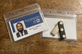 Two clear plastic ID badge holders on a wooden tabletop. The left holder contains a printed patient ID card, and the right holder contains a small black-and-silver folding knife with three round silver rivets.